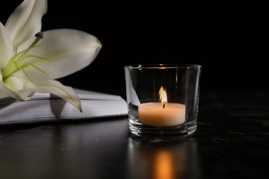 Burning candle, book and white lily on table in darkness. Funeral symbol