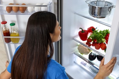 Young woman looking into modern refrigerator, back view