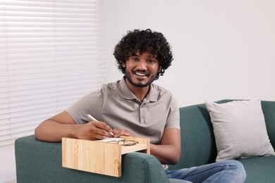 Happy man writing in notebook on sofa armrest wooden table at home