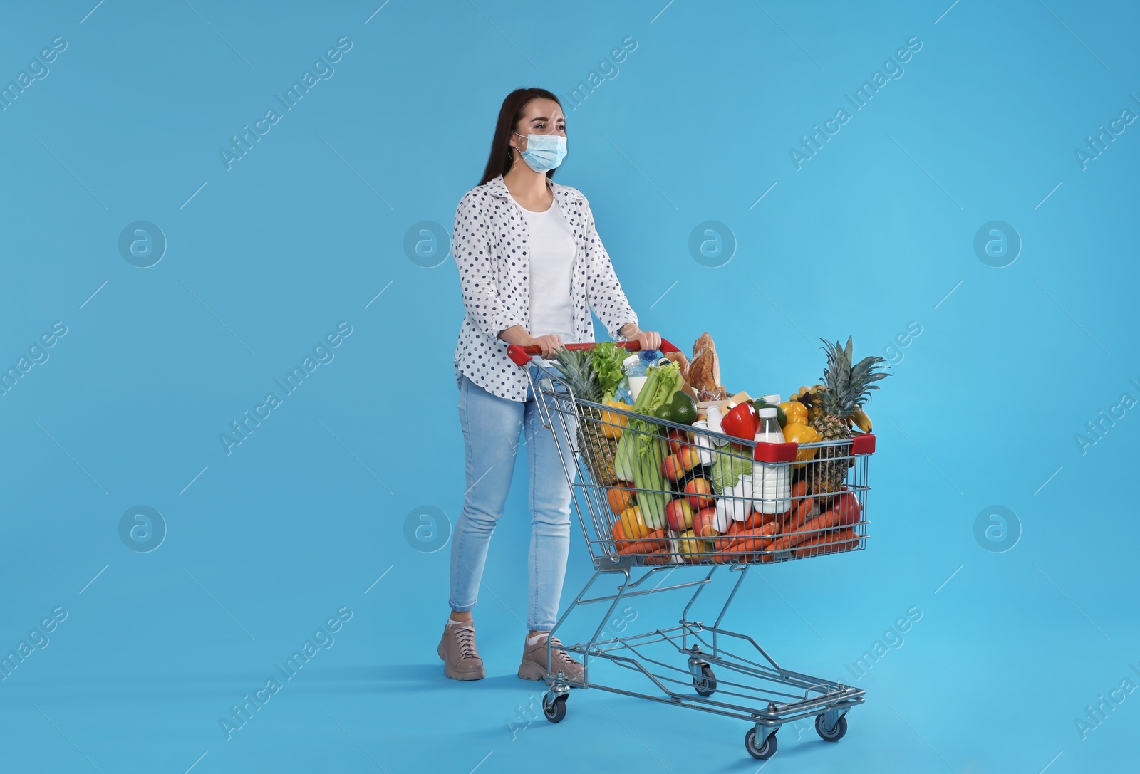 Photo of Young woman in medical mask with shopping cart full of groceries on light blue background