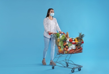 Young woman in medical mask with shopping cart full of groceries on light blue background