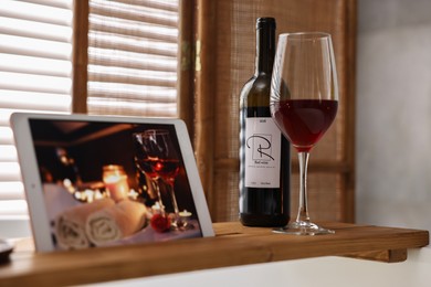 Photo of Wooden tray with tablet, glass of wine and bottle on bathtub in bathroom