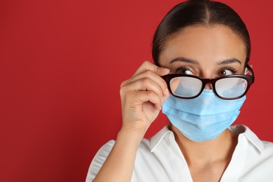 Photo of Woman wiping foggy glasses caused by wearing medical mask on red background. Space for text