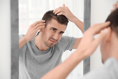 Photo of Young man with hair loss problem looking in mirror indoors