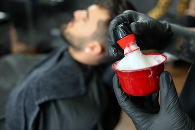 Photo of Professional hairdresser with shaving foam near bearded client in barbershop, closeup