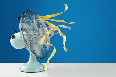 Photo of Electric fan on white table against blue background, space for text. Summer heat