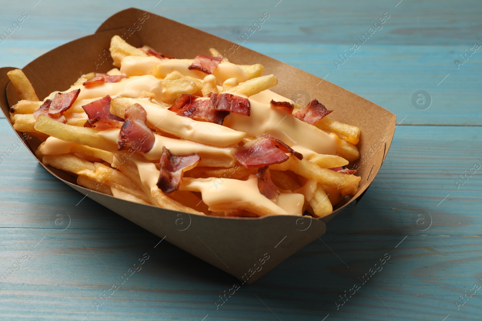 Photo of Tasty potato fries, cheese sauce and bacon in paper container on light blue wooden table, closeup