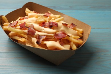 Photo of Tasty potato fries, cheese sauce and bacon in paper container on light blue wooden table, closeup