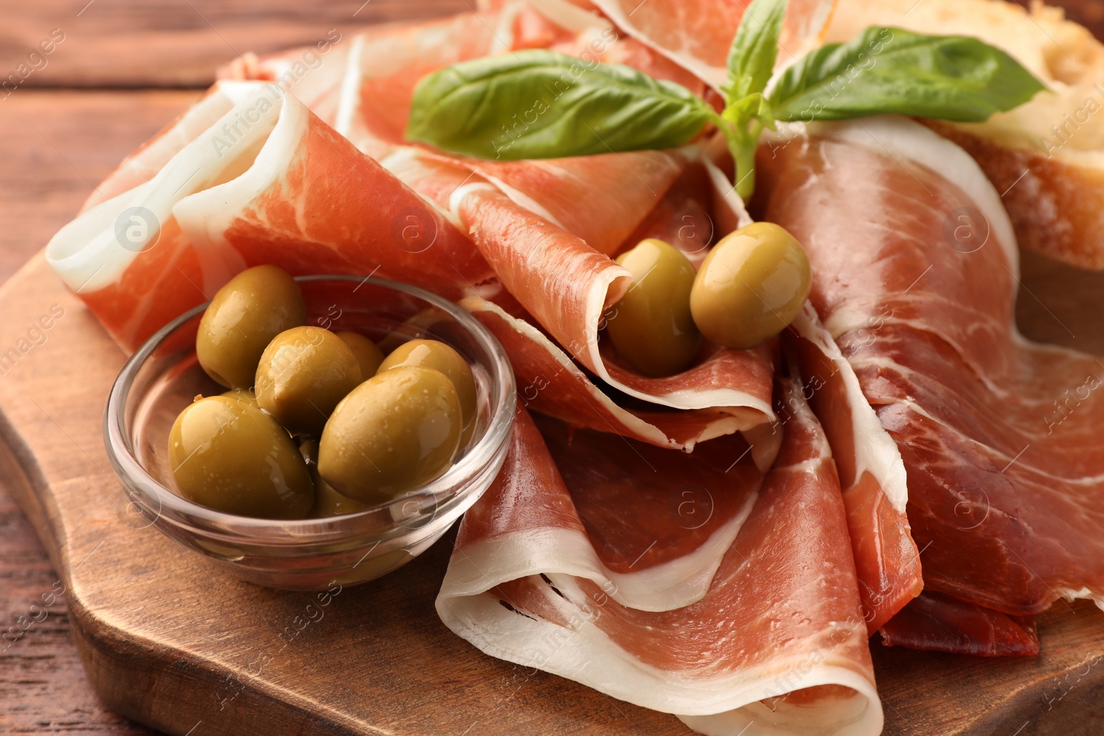 Photo of Slices of tasty cured ham, olives and basil on wooden board, closeup
