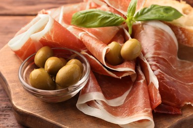 Photo of Slices of tasty cured ham, olives and basil on wooden board, closeup