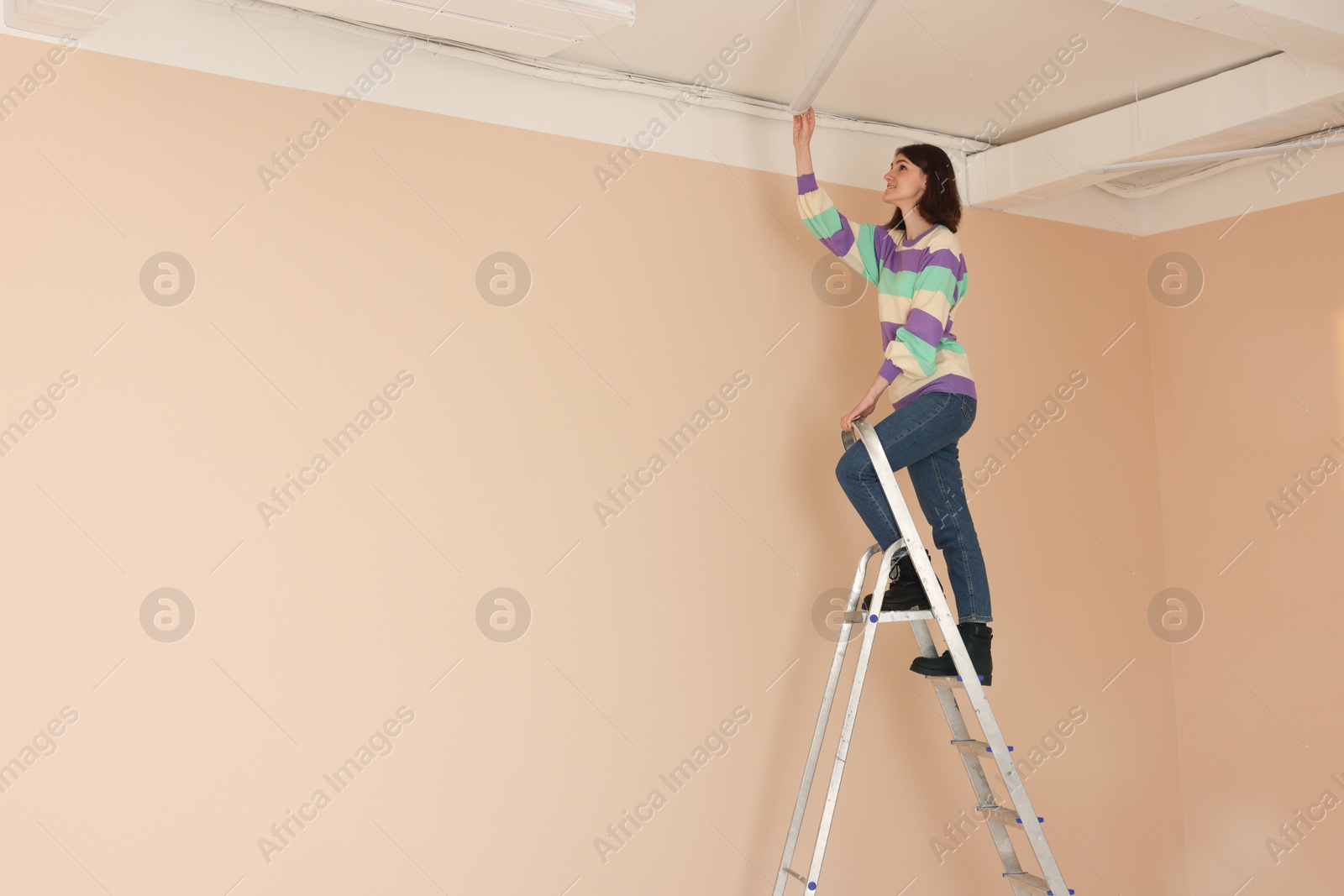 Photo of Young woman installing ceiling lamp on stepladder indoors, space for text