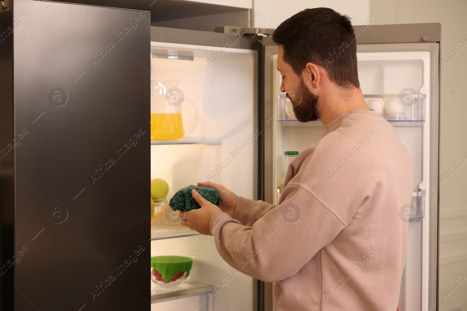 Photo of Man putting bowl covered with beeswax food wrap into refrigerator indoors