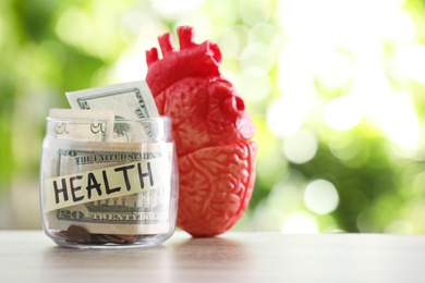 Glass jar with money and heart model on table against blurred background. Space for text