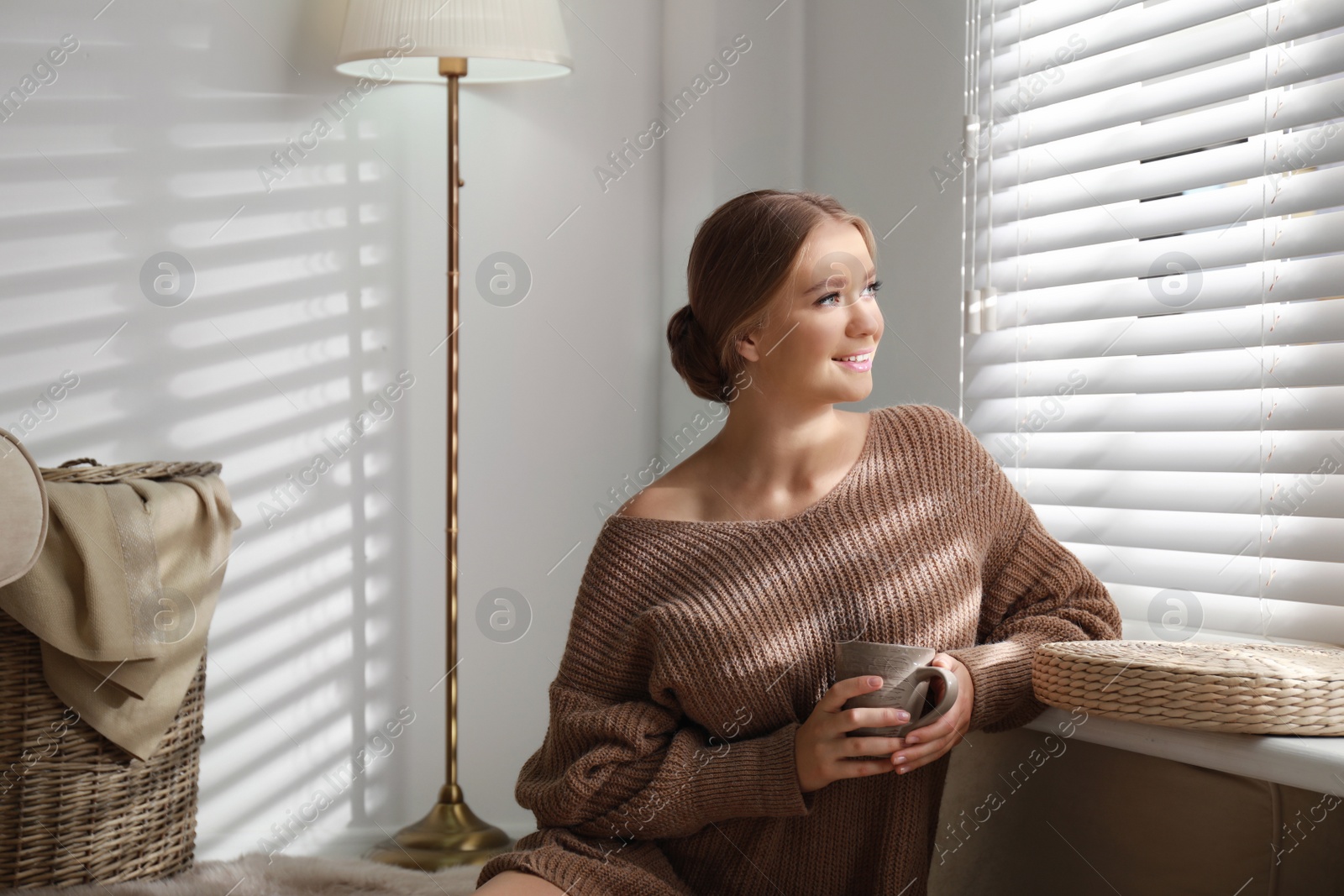 Photo of Beautiful young woman with cup of hot drink near window at home. Winter atmosphere