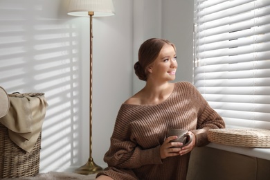 Photo of Beautiful young woman with cup of hot drink near window at home. Winter atmosphere