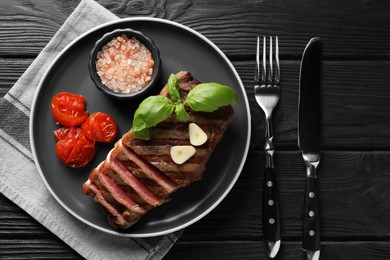Delicious grilled beef steak served with spices and tomatoes on black wooden table, flat lay