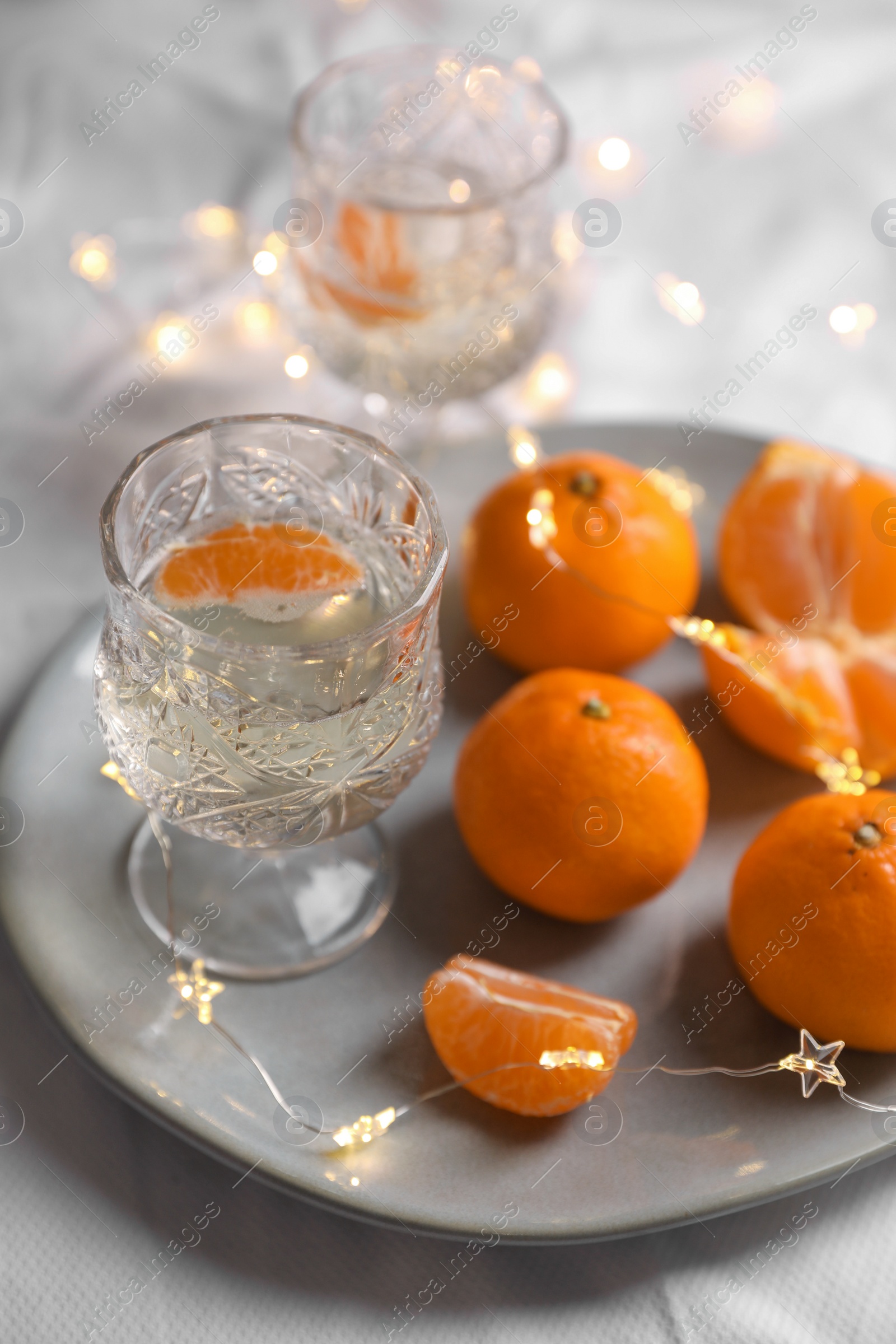 Photo of Delicious ripe tangerines, glasses of sparkling wine and fairy lights on white bedsheet