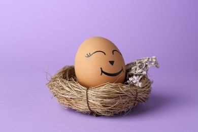 Photo of Egg with drawn happy face in nest on lilac background, closeup