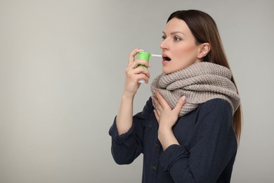 Woman with scarf using throat spray on grey background, space for text