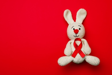 Cute knitted toy bunny with ribbon on red background, top view and space for text. AIDS disease awareness