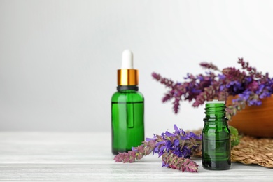 Photo of Bottles of sage essential oil and flowers on white table, space for text