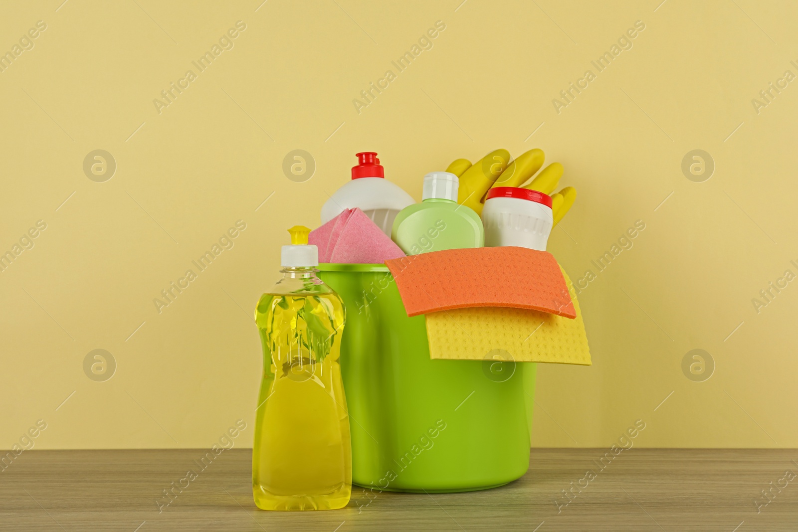 Photo of Bucket with different cleaning supplies on wooden floor near beige wall