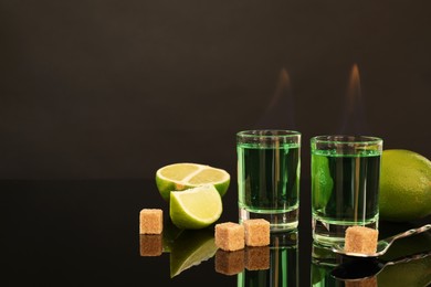 Photo of Flaming absinthe in shot glasses, brown sugar, lime and spoon on mirror table, space for text. Alcoholic drink