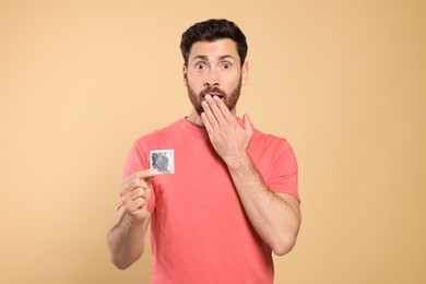 Emotional man holding condom on beige background. Safe sex