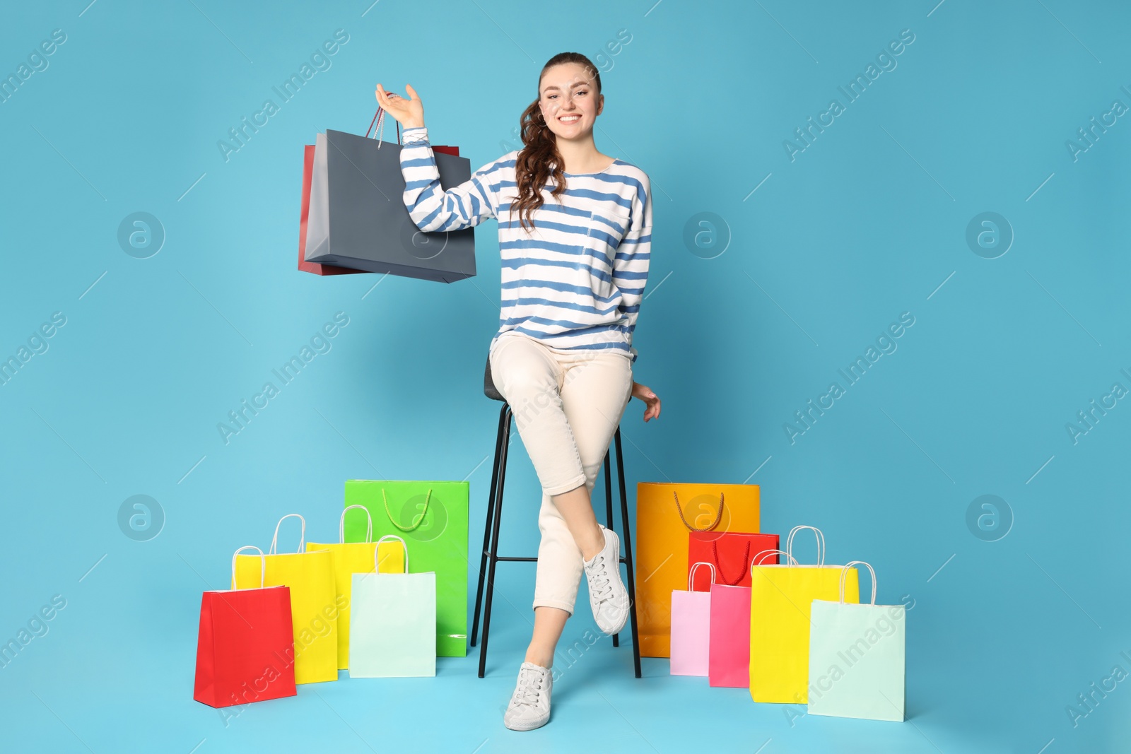 Photo of Happy woman holding colorful shopping bags on stool against light blue background