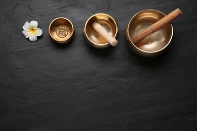 Photo of Golden singing bowls, mallets and flower on black table, flat lay. Space for text