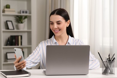 Photo of Happy woman with smartphone working on laptop at white desk in room