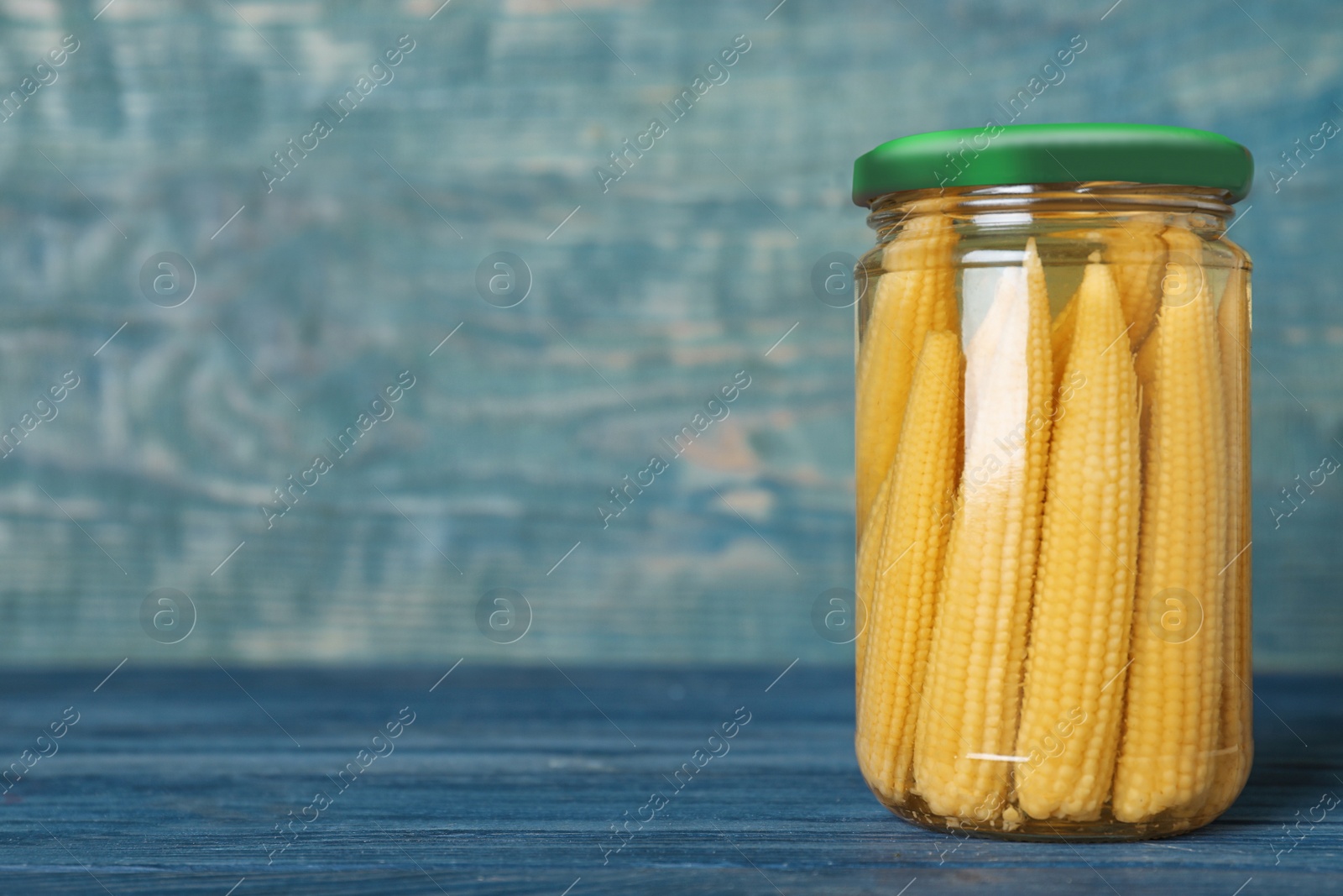 Photo of Glass jar of pickled corn cobs on blue wooden table. Space for text