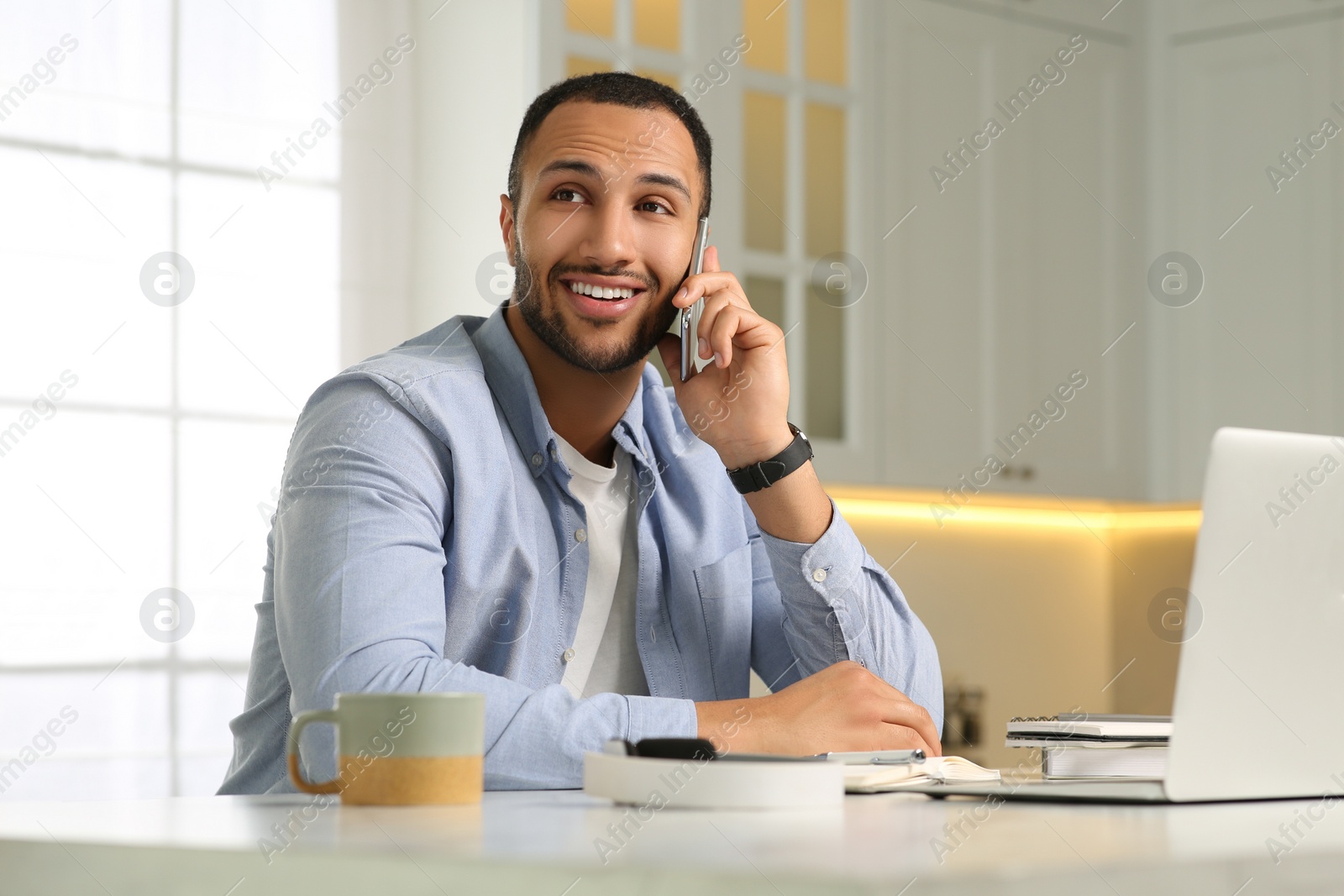 Photo of Young man talking on smartphone while working with laptop at desk in kitchen. Home office