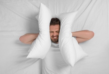 Photo of Irritated man covering his ears with pillows in bed, top view. Insomnia problem