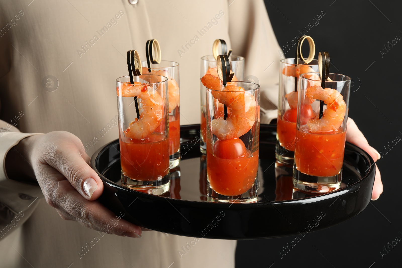 Photo of Woman holding tasty canapes with shrimps, tomatoes and sauce in shot glasses on black background, closeup