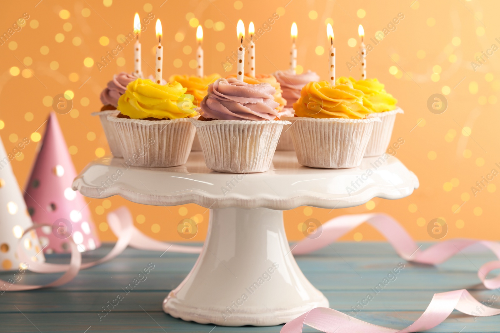 Photo of White stand with tasty birthday cupcakes on light blue wooden table against blurred lights