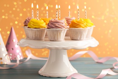 Photo of White stand with tasty birthday cupcakes on light blue wooden table against blurred lights