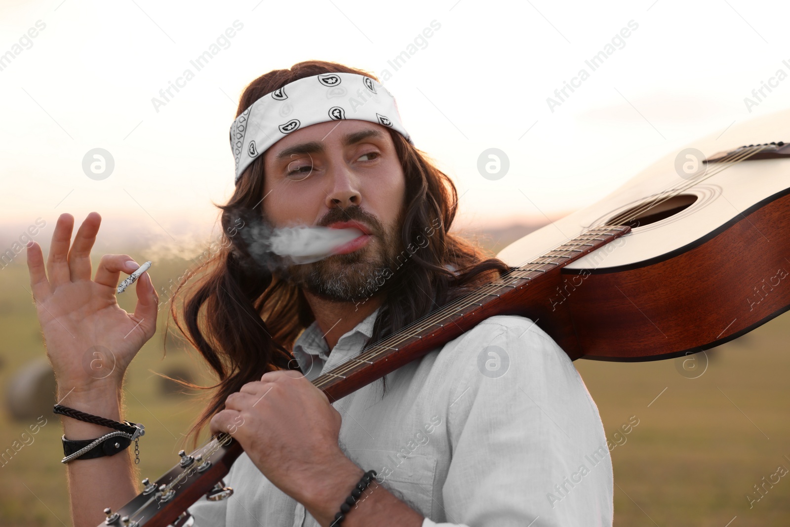 Photo of Stylish hippie man with guitar smoking joint in field