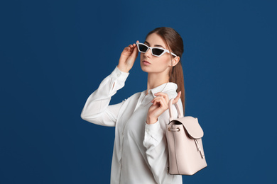 Photo of Beautiful young woman in sunglasses with stylish bag on blue background