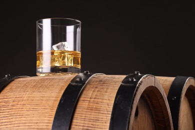 Whiskey with ice cubes in glass on wooden barrel against dark background, space for text