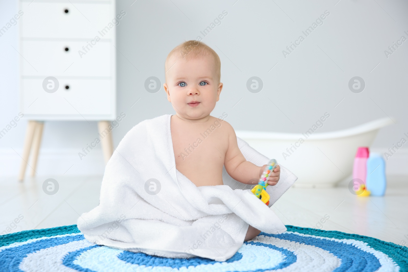 Photo of Cute little baby with soft towel and rattle on rug in bathroom