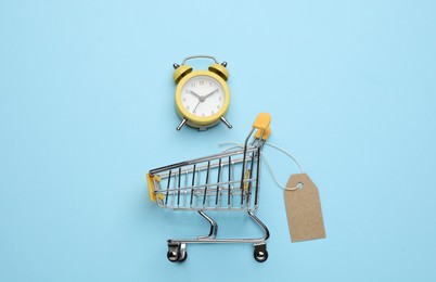 Photo of Shopping cart with tag and alarm clock on light blue background, flat lay. Sale concept