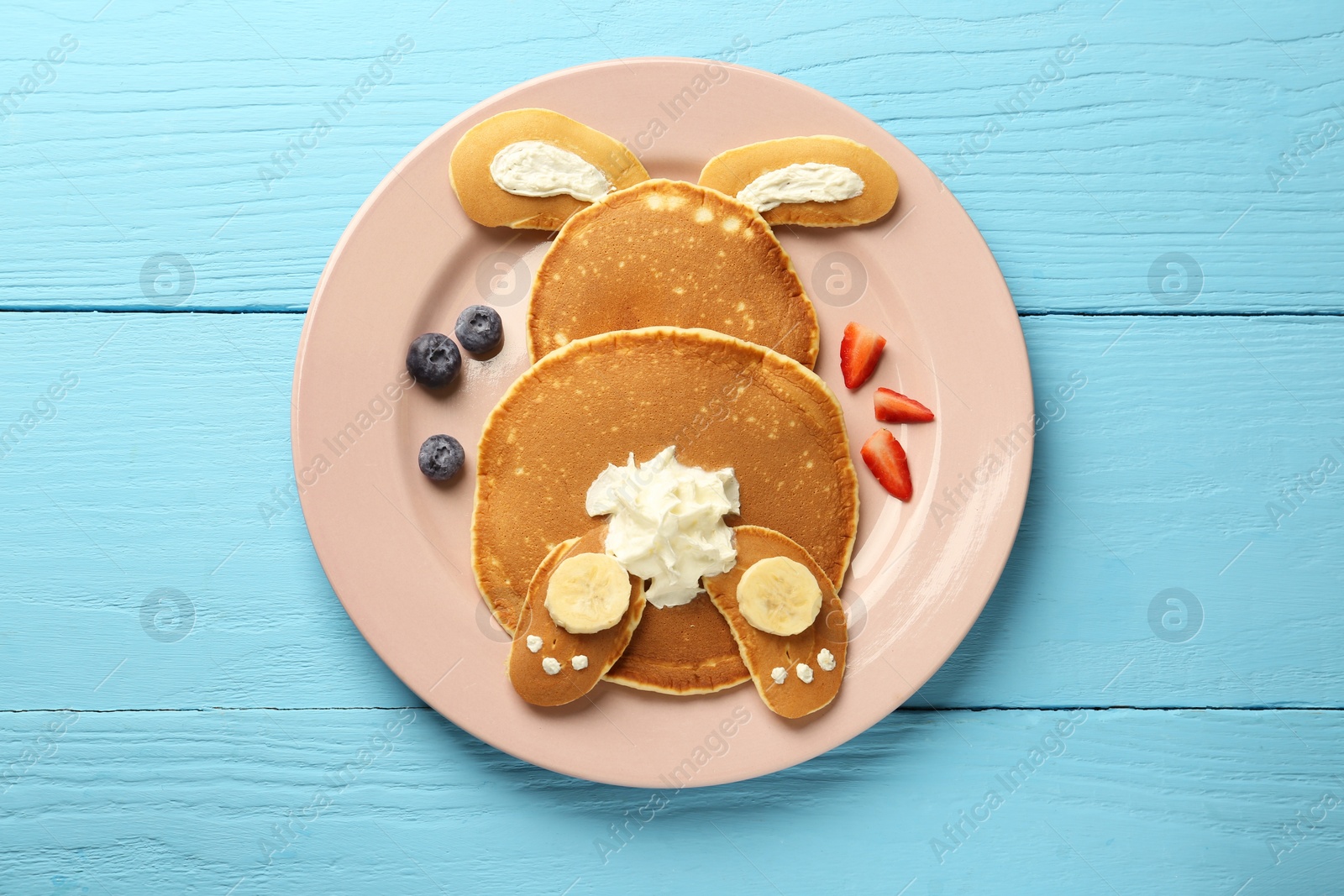 Photo of Creative serving for kids. Plate with cute bunny made of pancakes, berries, cream and banana on light blue wooden table, top view