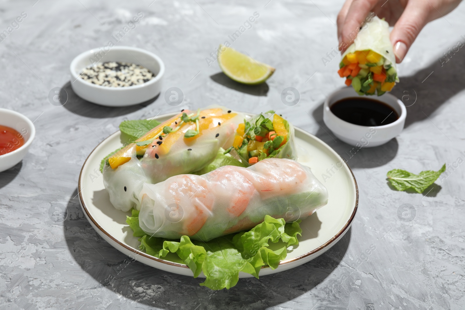 Photo of Plate with tasty spring rolls on grey textured table. Woman dipping delicious roll into soy sauce, closeup