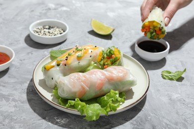 Plate with tasty spring rolls on grey textured table. Woman dipping delicious roll into soy sauce, closeup