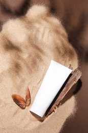 Photo of Tube of body cream, seashell and tree bark on sand against brown background, flat lay