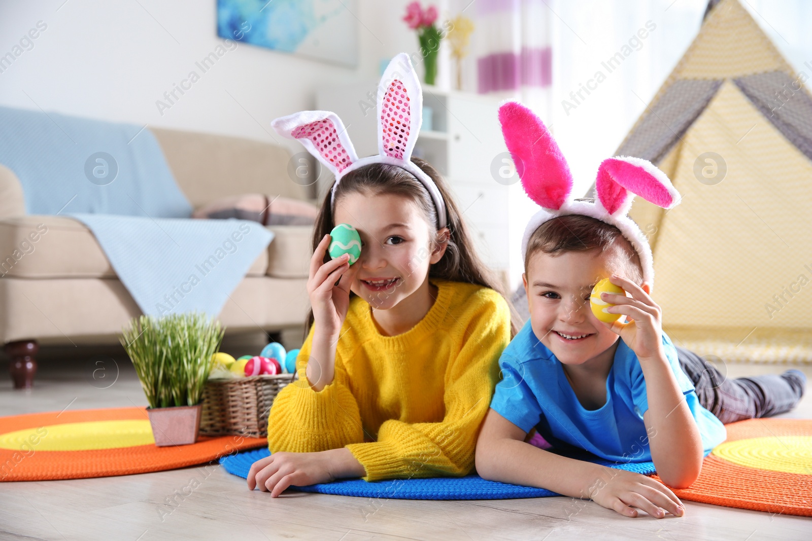 Photo of Cute children with bunny headbands and painted Easter eggs lying on floor at home