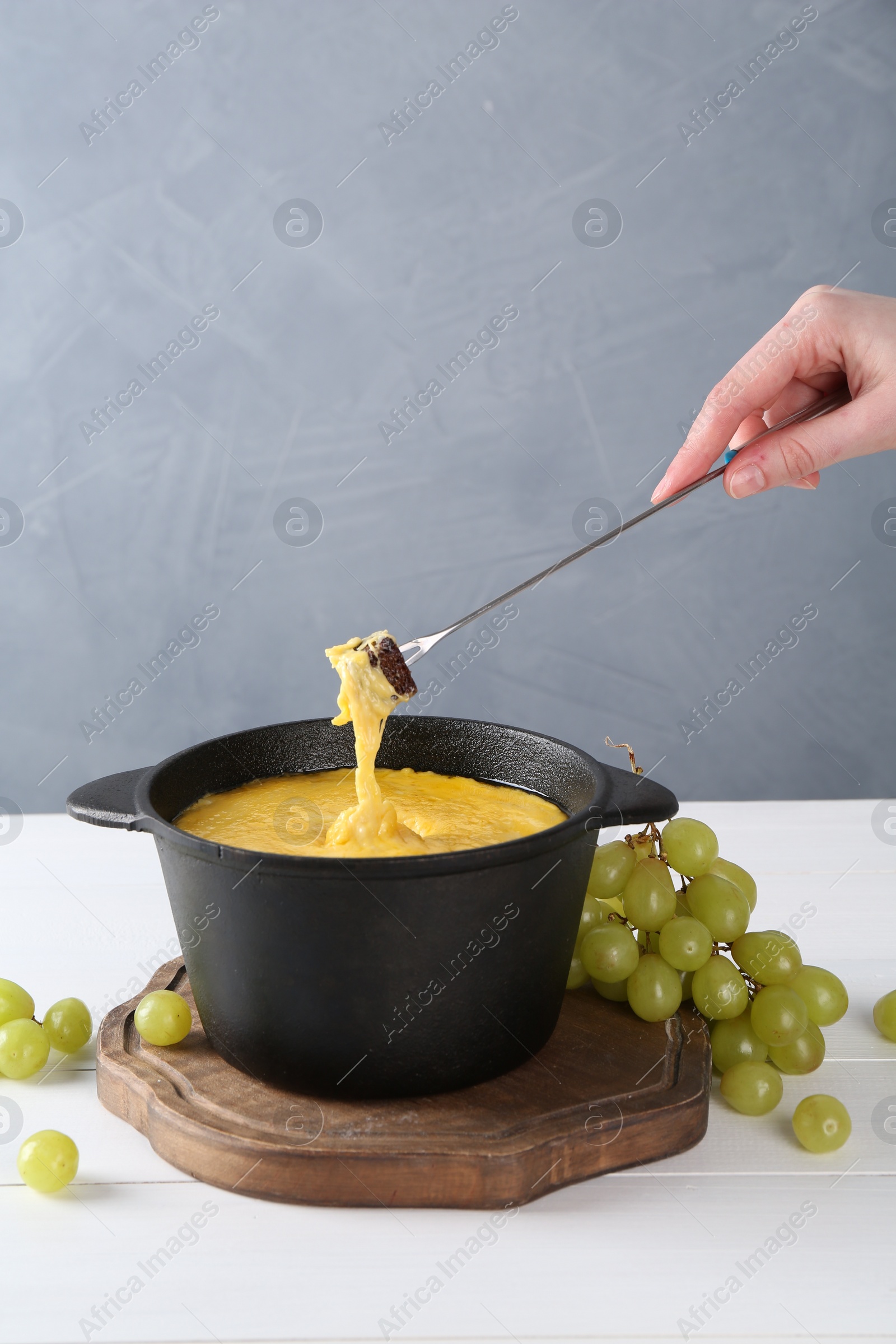 Photo of Woman dipping piece of bread into fondue pot with tasty melted cheese at white wooden table against gray background, closeup