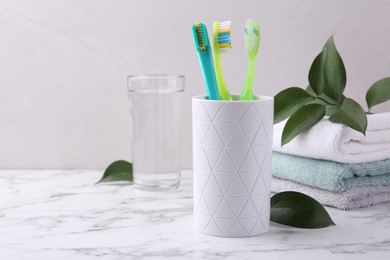 Photo of Colorful plastic toothbrushes in container, towels and green leaves on white marble table, space for text