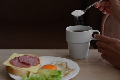 Woman adding sugar to morning coffee while having breakfast at wooden table indoors, closeup